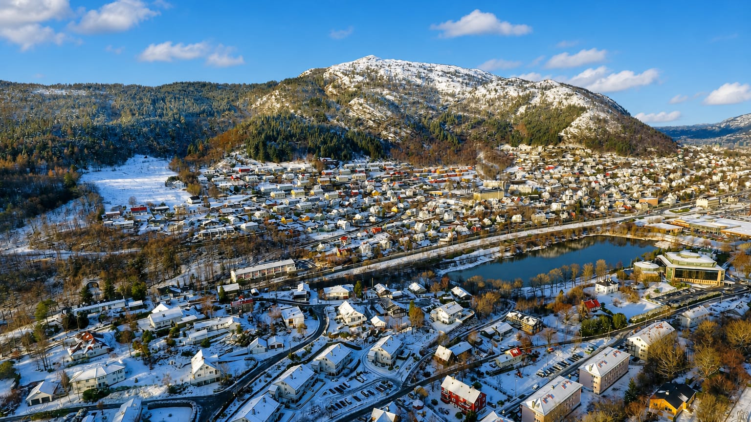 Snow-covered residential neighborhood of Minde, Norway, beside a small lake below a forested mountain under a blue sky.