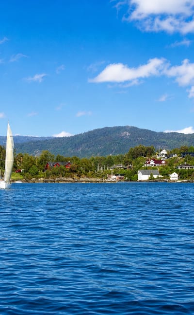 Sailboat on the fjord at Ytrebygda, Bergen, with wooded shores, scattered houses and distant mountains under a blue sky.