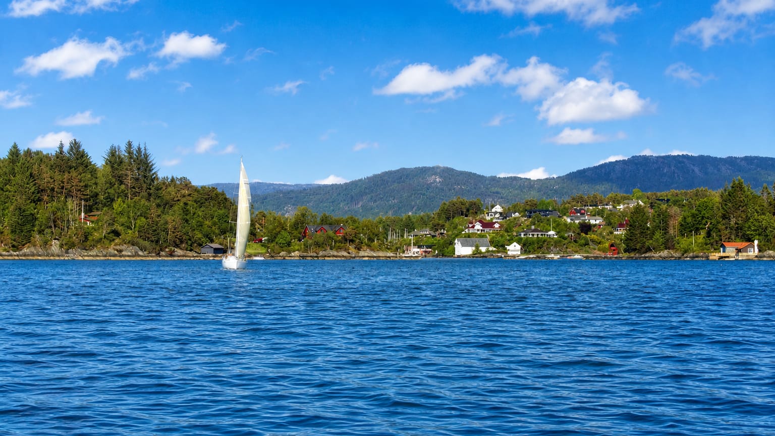 Sailboat on the fjord at Ytrebygda, Bergen, with wooded shores, scattered houses and distant mountains under a blue sky.