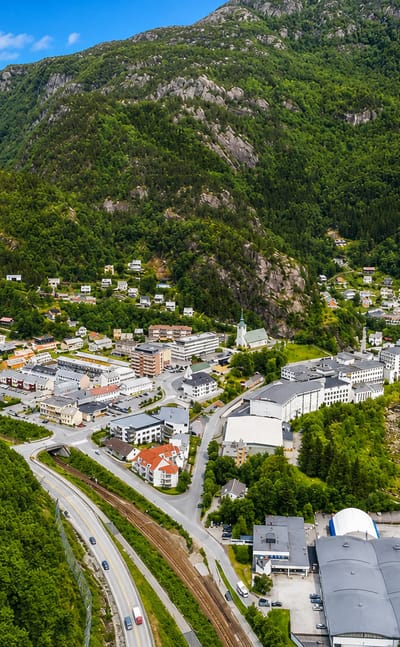 Aerial view of Dale in Vaksdal, Norway, a valley village with industrial buildings and homes below steep forested mountains.