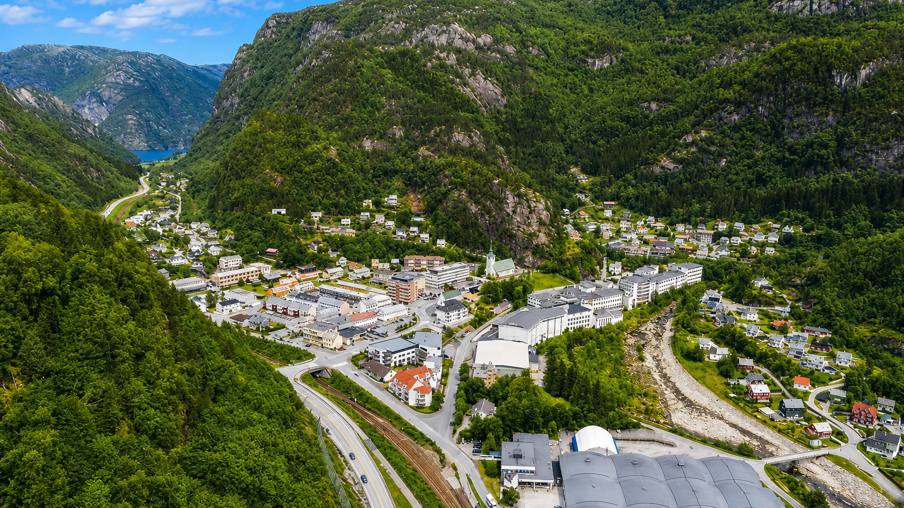Aerial view of Dale in Vaksdal, Norway, a valley village with industrial buildings and homes below steep forested mountains.