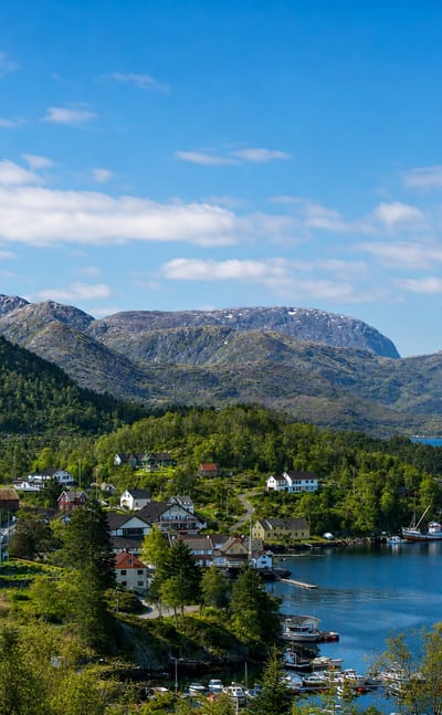 Eivindvik's white wooden church above a calm fjord, ringed by forested mountains and scattered houses under a blue sky.