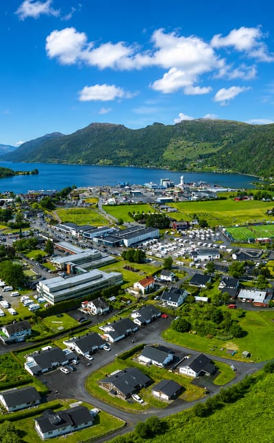 Aerial view of Etne, Norway, a fjordside village of white houses among green farmland with a river beneath forested mountains.