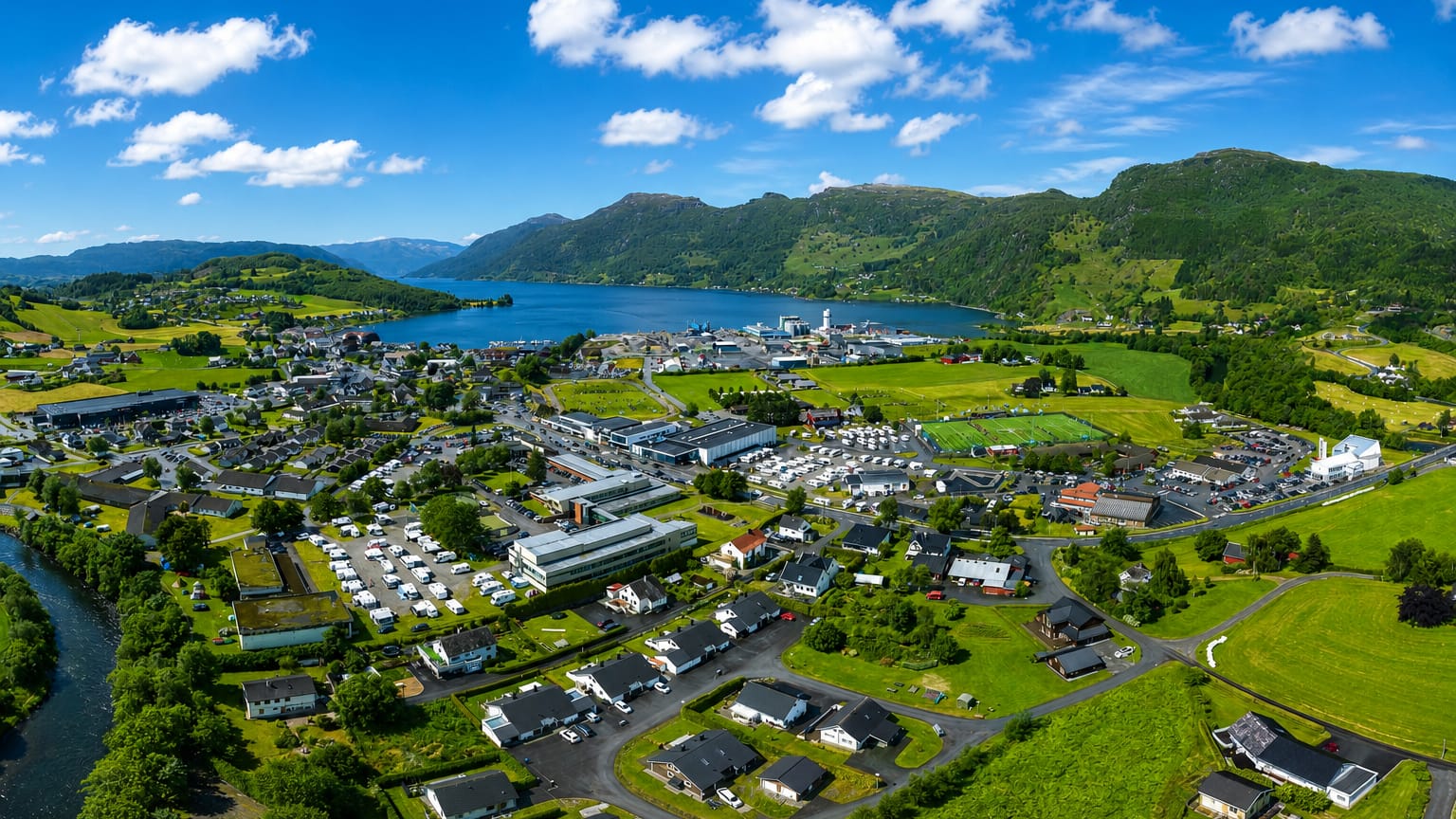 Aerial view of Etne, Norway, a fjordside village of white houses among green farmland with a river beneath forested mountains.