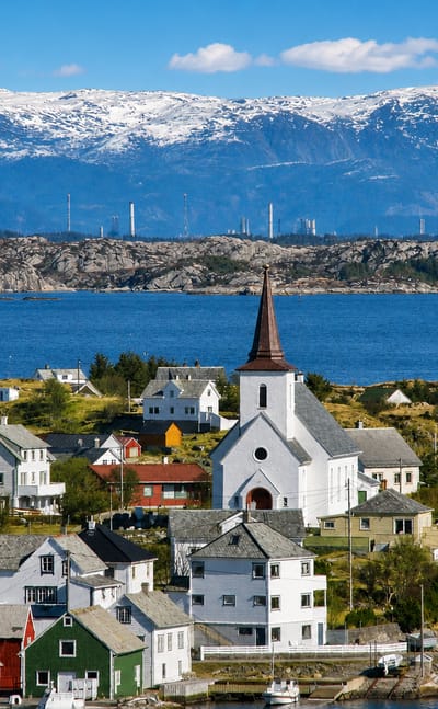 Colorful wooden houses and a white steepled church by the sea in Fedje, Norway, with distant snow-capped mountains.