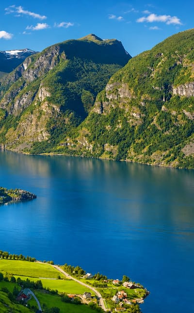 Flåm village beside the deep blue Aurlandsfjord, framed by steep green mountains and snow-flecked peaks under a bright sky.