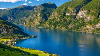 Flåm village beside the deep blue Aurlandsfjord, framed by steep green mountains and snow-flecked peaks under a bright sky.