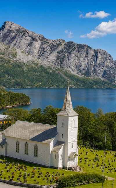 White wooden church and fjord-side cemetery in Hyllestad, Norway, with steep mountains rising across the calm fjord.