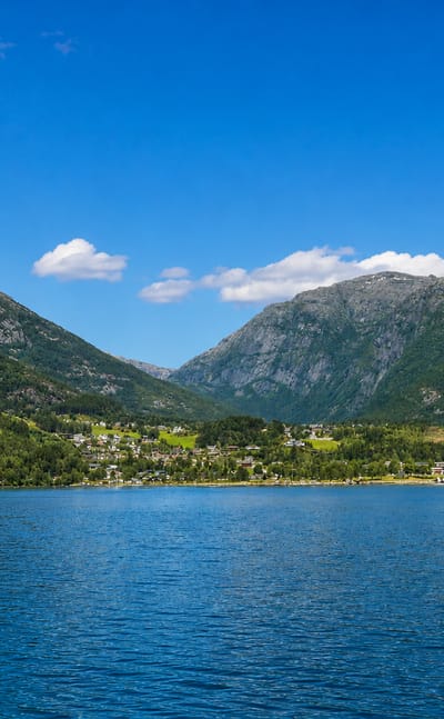 Kinsarvik village on the shore of the Sørfjorden, nestled beneath steep forested mountains under a clear blue sky.