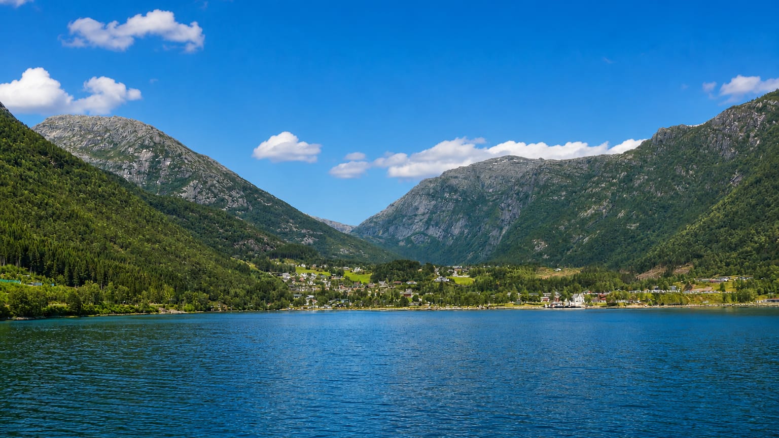 Kinsarvik village on the shore of the Sørfjorden, nestled beneath steep forested mountains under a clear blue sky.