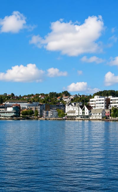 Leirvik waterfront in Norway across calm water, with white gabled quayside buildings and a tall lattice communications tower on the wooded hill.