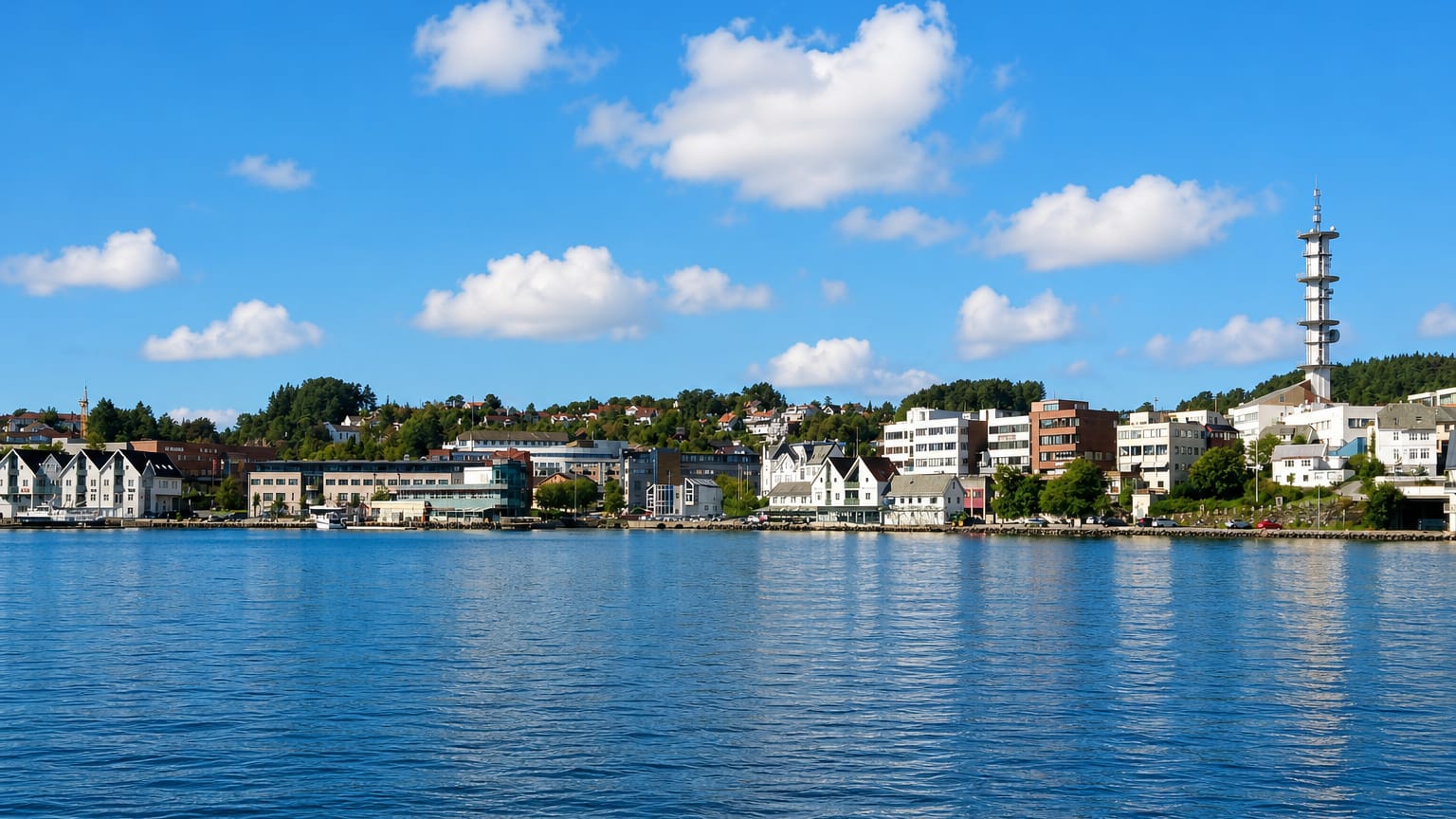Leirvik waterfront in Norway across calm water, with white gabled quayside buildings and a tall lattice communications tower on the wooded hill.