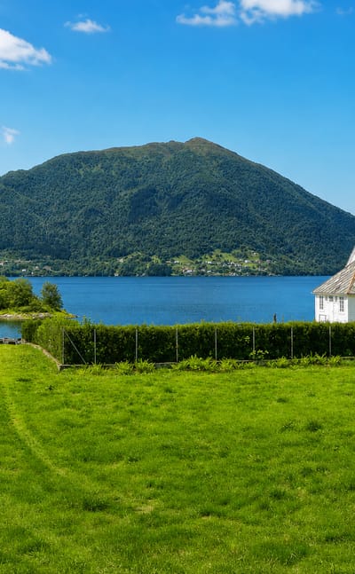 A white timber church beside a calm fjord with a green meadow and forested mountains near Lonevåg, Norway.