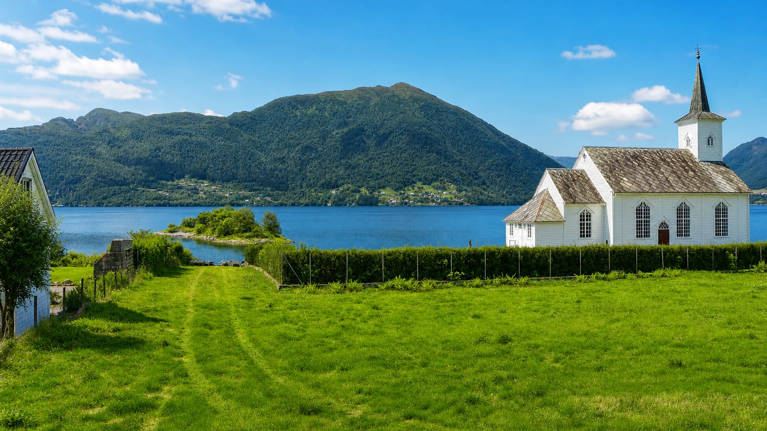 A white timber church beside a calm fjord with a green meadow and forested mountains near Lonevåg, Norway.