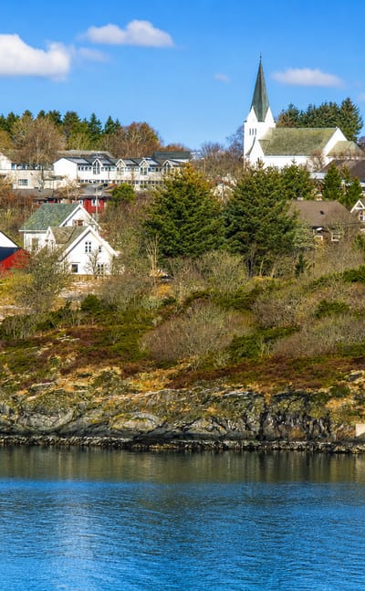 The waterfront village of Manger, Norway, its white spired church rising above wooden houses across a calm blue fjord.