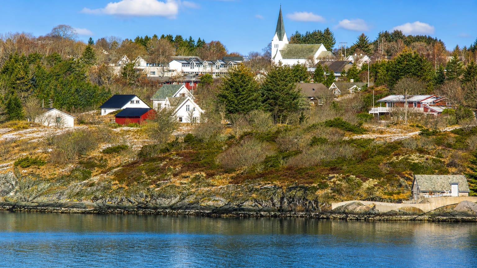 The waterfront village of Manger, Norway, its white spired church rising above wooden houses across a calm blue fjord.