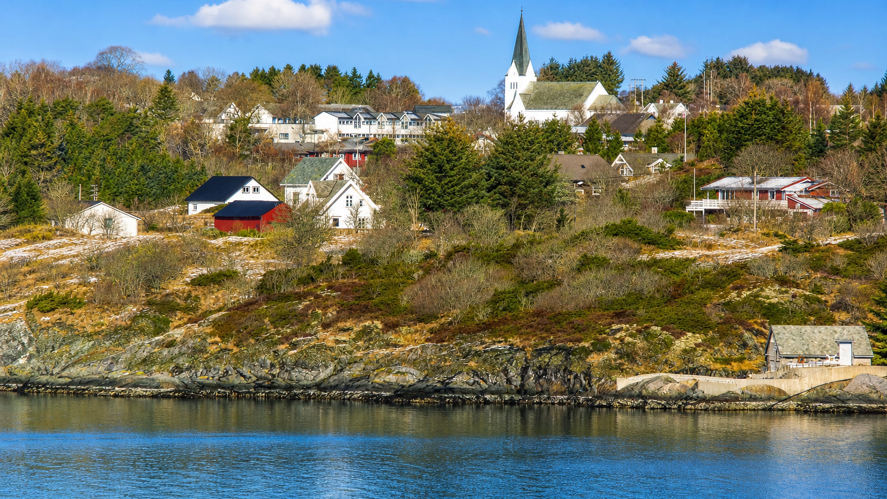The waterfront village of Manger, Norway, its white spired church rising above wooden houses across a calm blue fjord.