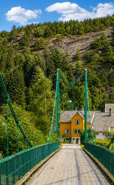 Green suspension footbridge to Mo's white wooden church and yellow houses below a forested hillside, Norway