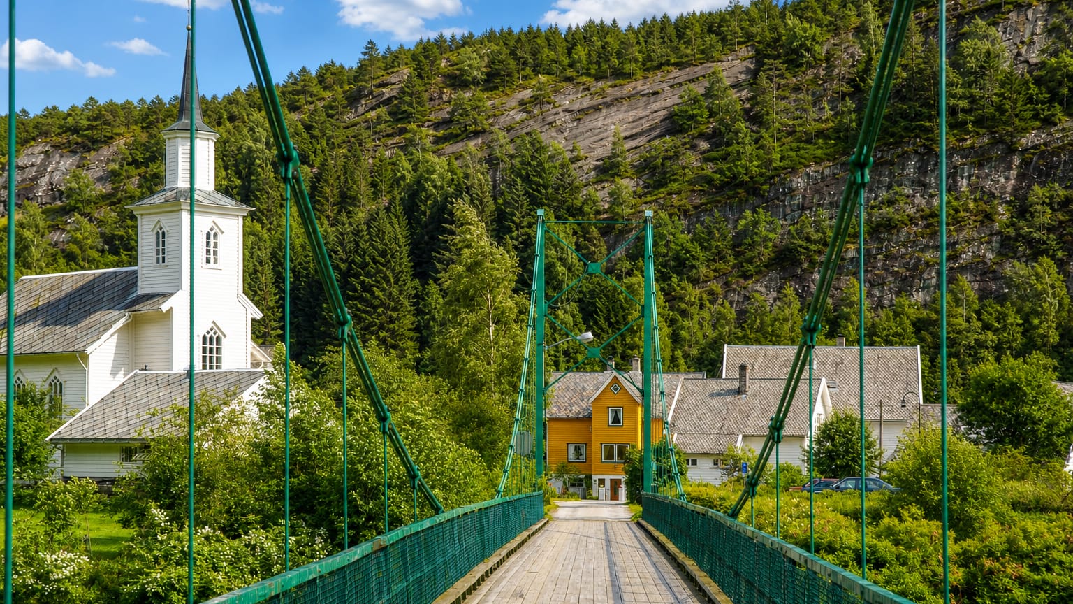 Green suspension footbridge to Mo's white wooden church and yellow houses below a forested hillside, Norway