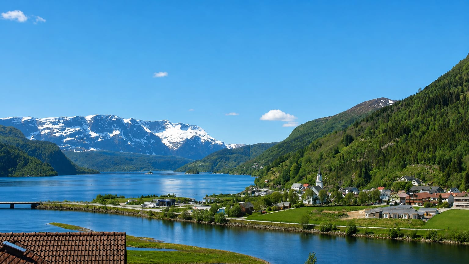 Naustdal, Norway, a fjord village with a white church beside blue water below snow-capped Vestland mountains.