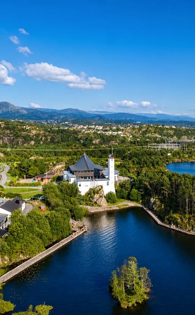 Aerial view of Straume, Norway: a white tower villa and pool beside forested lakes with the town beyond low hills