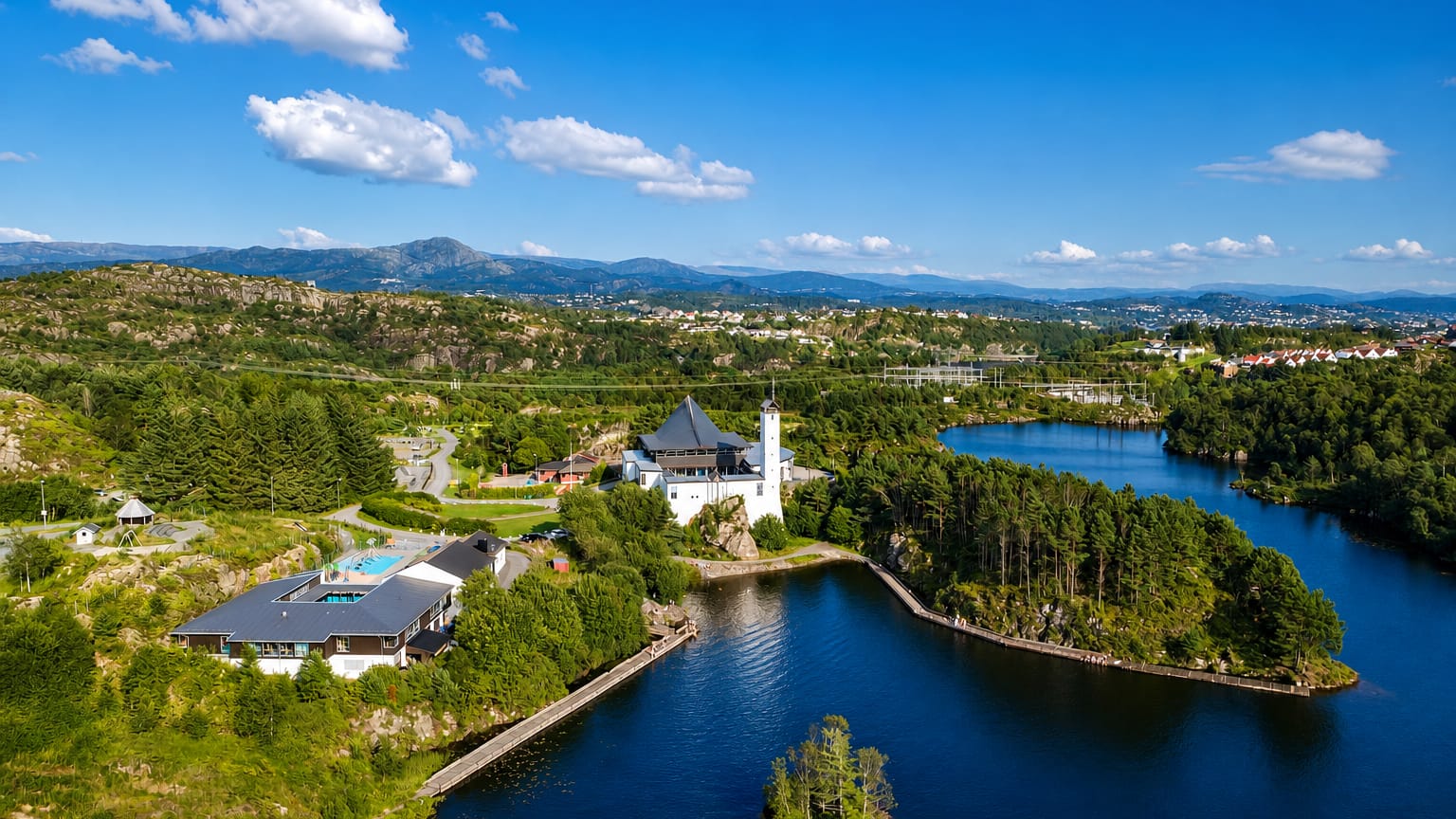 Aerial view of Straume, Norway: a white tower villa and pool beside forested lakes with the town beyond low hills