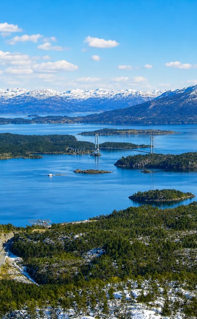 Suspension bridge spanning a fjord among wooded islands with snow-capped mountains at Sveio