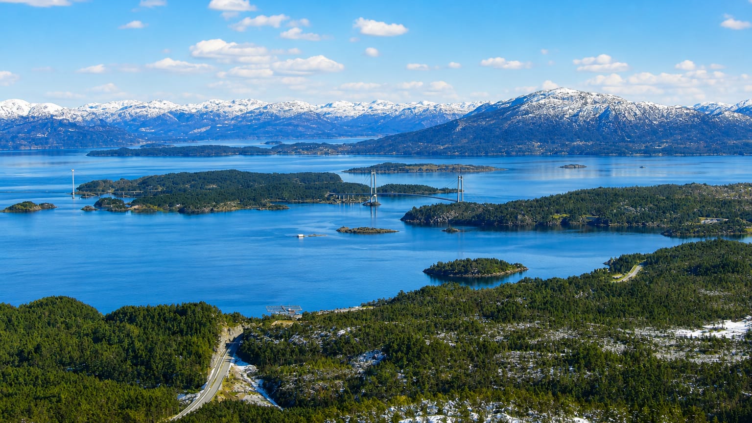 Suspension bridge spanning a fjord among wooded islands with snow-capped mountains at Sveio