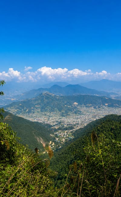 Kathmandu Valley and city sprawl viewed from a forested hillside with Himalayan foothills beyond under blue sky