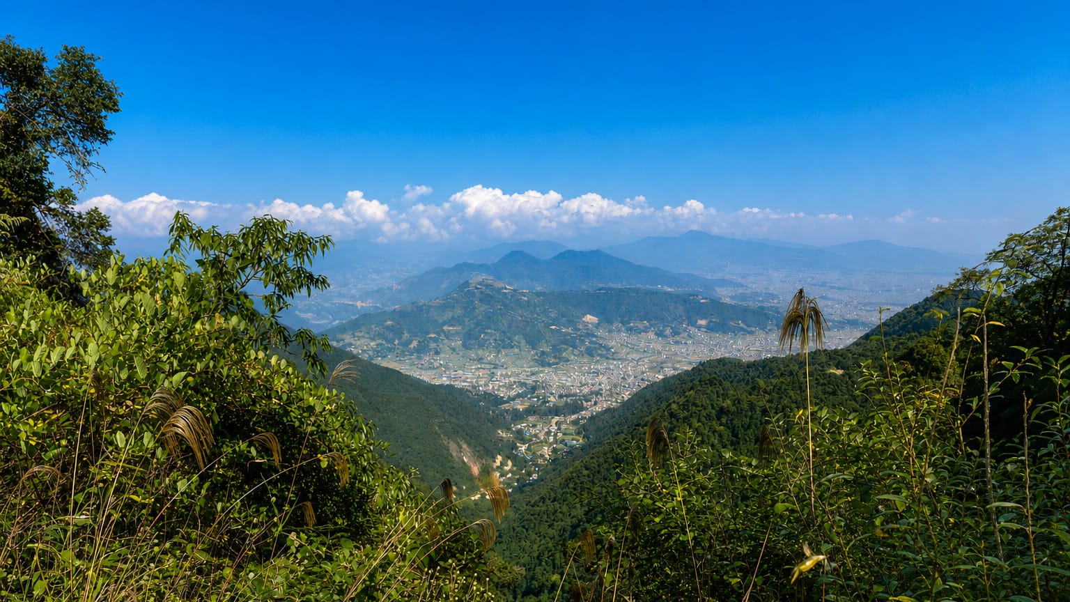 Kathmandu Valley and city sprawl viewed from a forested hillside with Himalayan foothills beyond under blue sky
