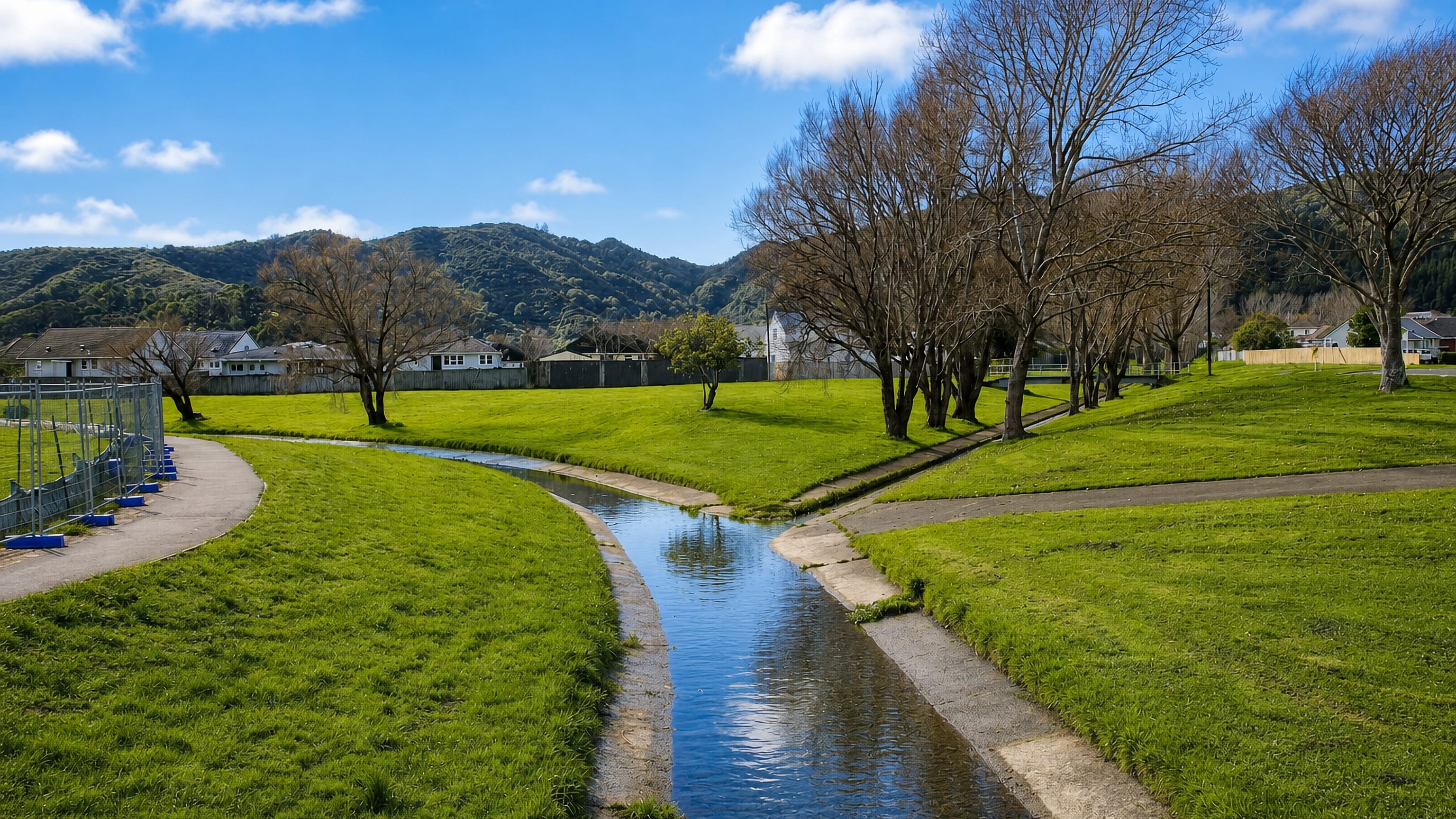 Grassy park with a channelled stream and bush-covered hills behind suburban houses in Naenae, Lower Hutt.