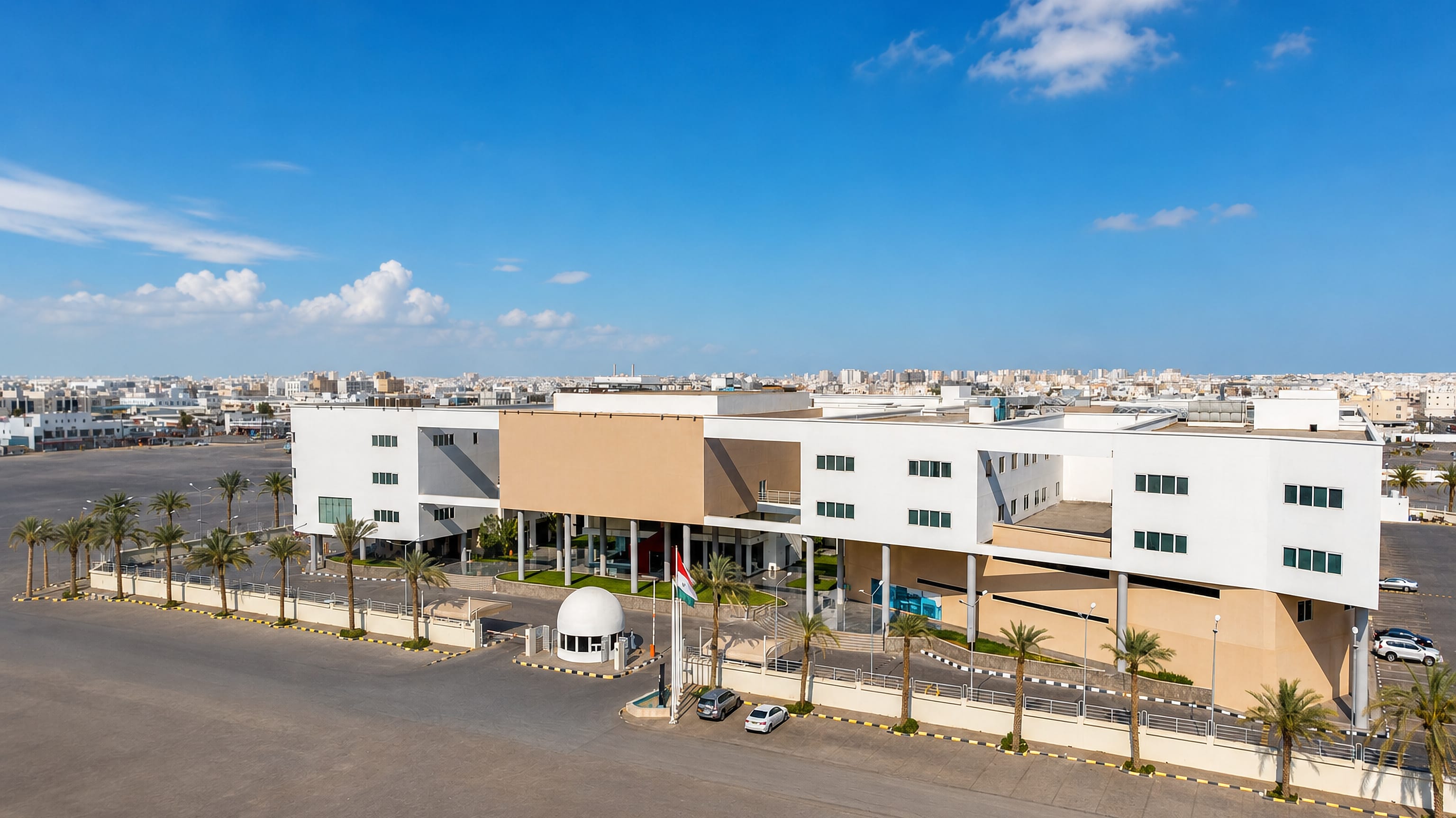 A modern white-and-beige civic building lined with palms in Seeb, Oman, with the low-rise town behind
