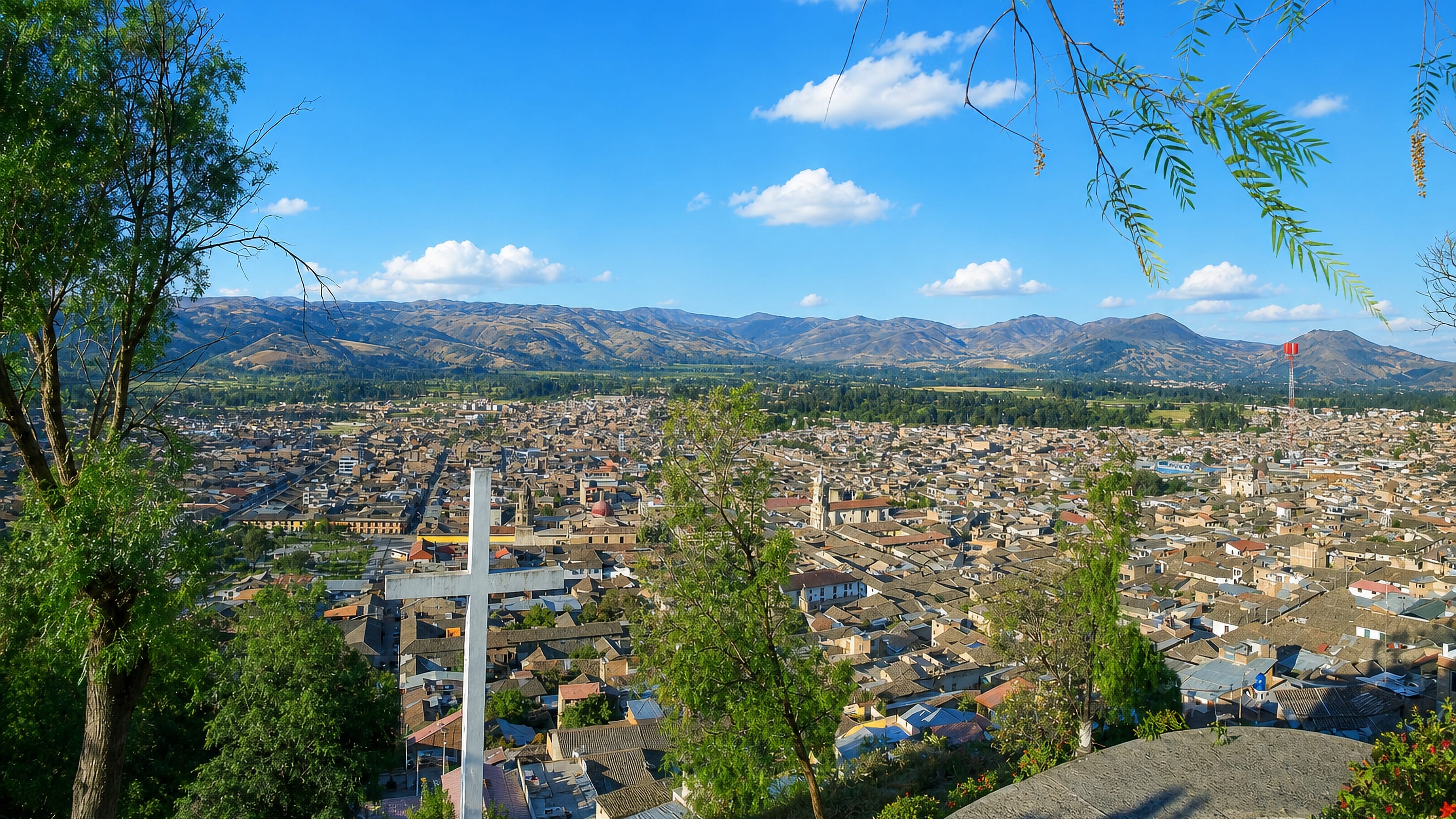 Hilltop view over Cajamarca, Peru, its valley rooftops and church towers framed by the Andes and a white cross.