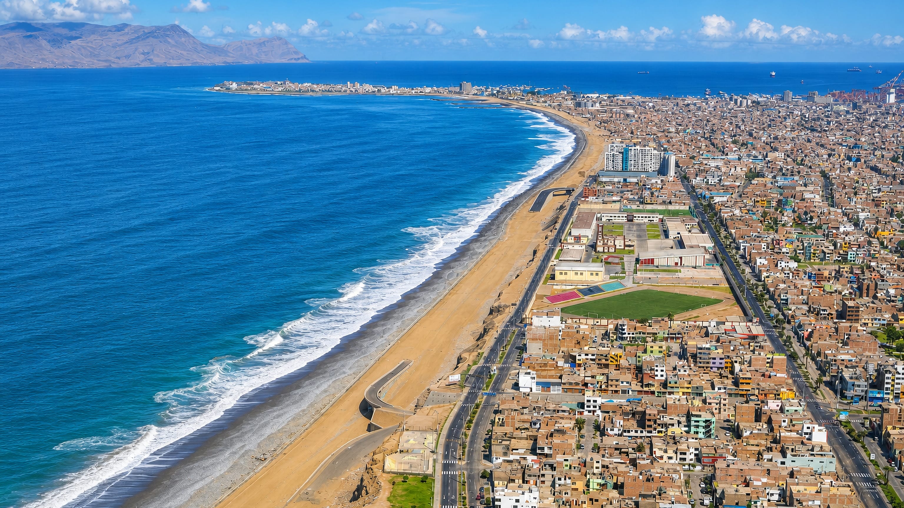 Aerial view of La Perla, Callao, a long sandy beach and coastal road beside dense low-rise blocks and a headland.