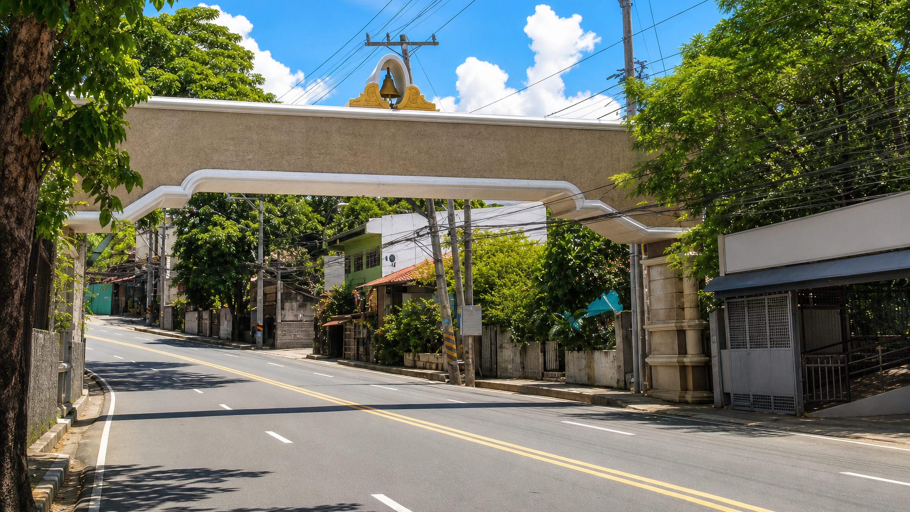A quiet road passes beneath a bell-topped concrete welcome arch on a tree-lined street in Antipolo, Philippines.