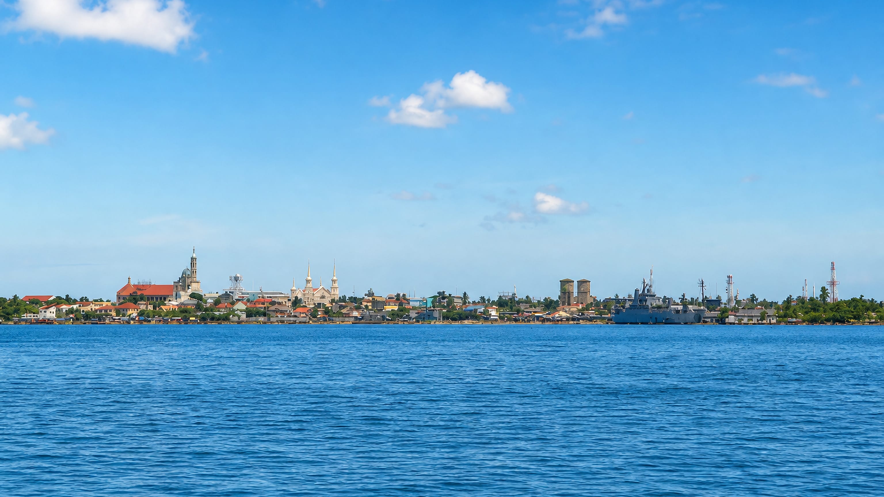 Cavite City's low waterfront of church spires and a naval pier seen across the blue water of Manila Bay.