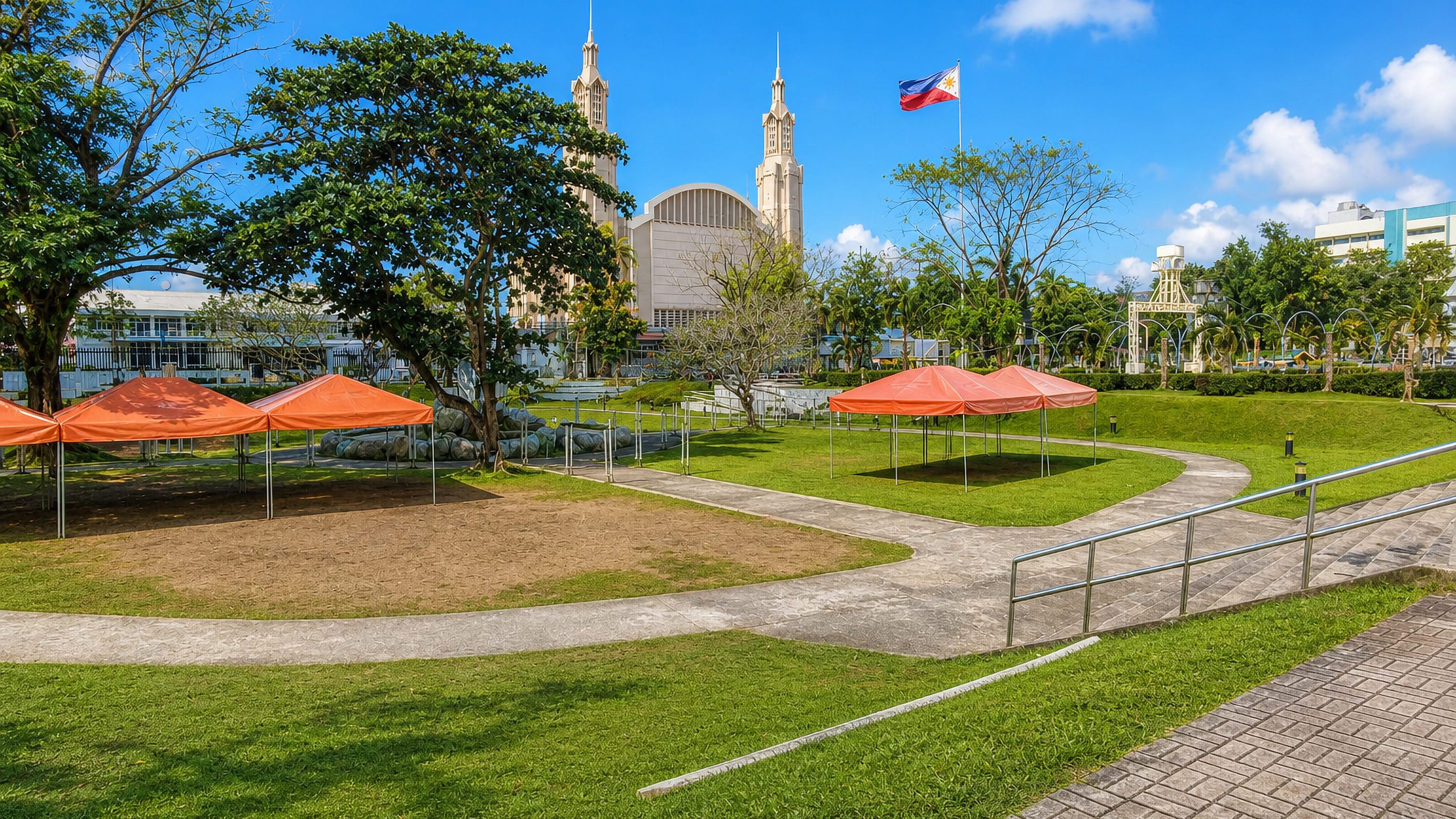 Park lawn with event canopies and the Philippine flag in front of a twin-spired church in Lucena, Philippines.