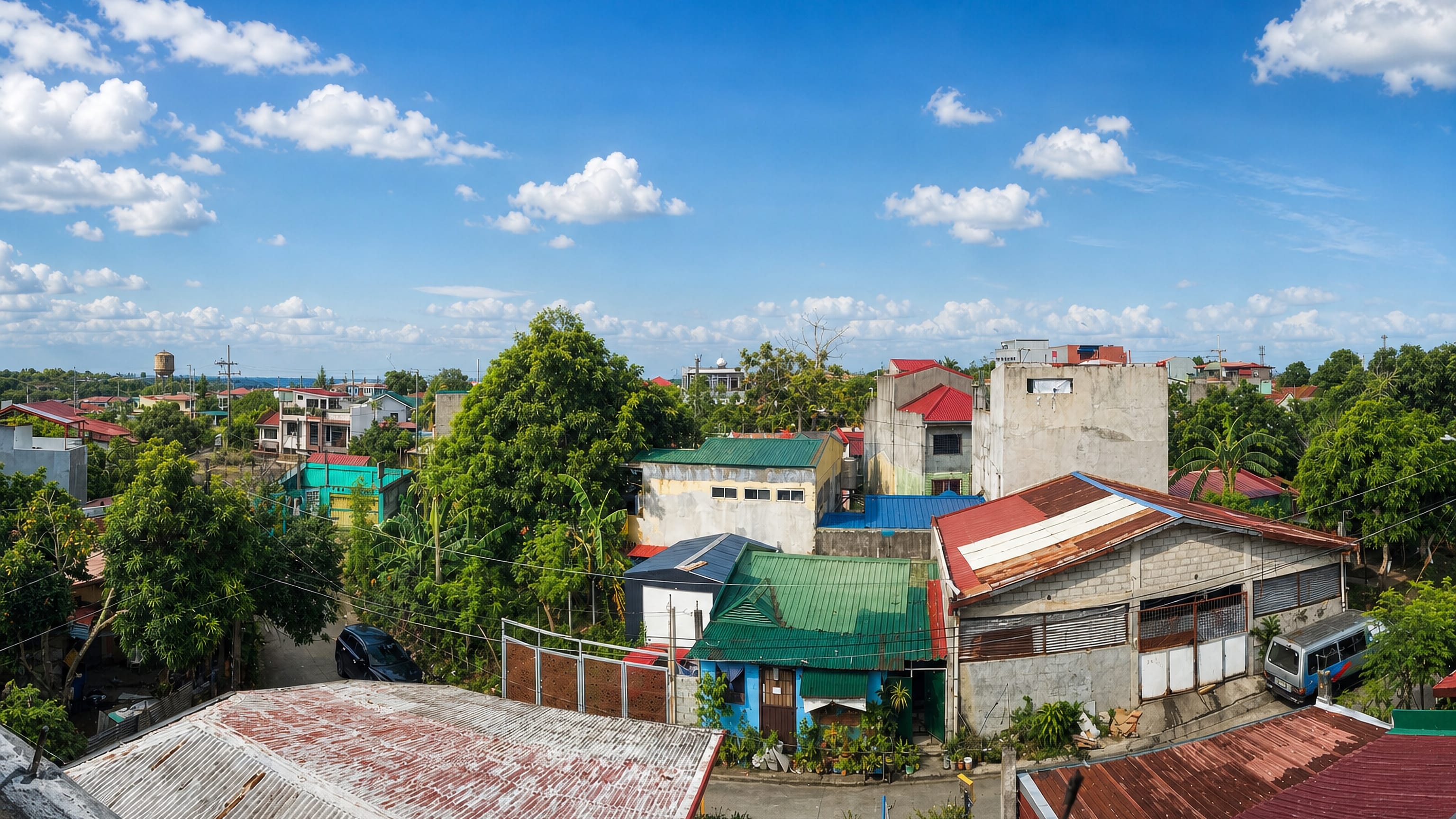 Elevated view over Silang, Philippines, with corrugated-roof houses, mango trees and a water tower under blue sky