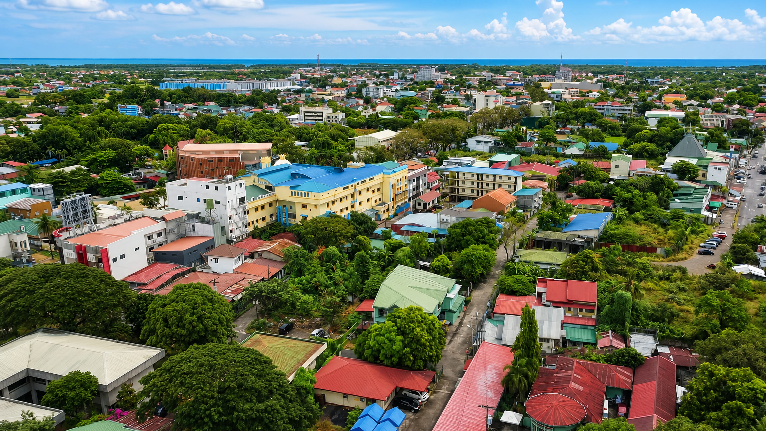 Aerial view over Balanga's colourful low-rise rooftops and trees with Manila Bay on the horizon.