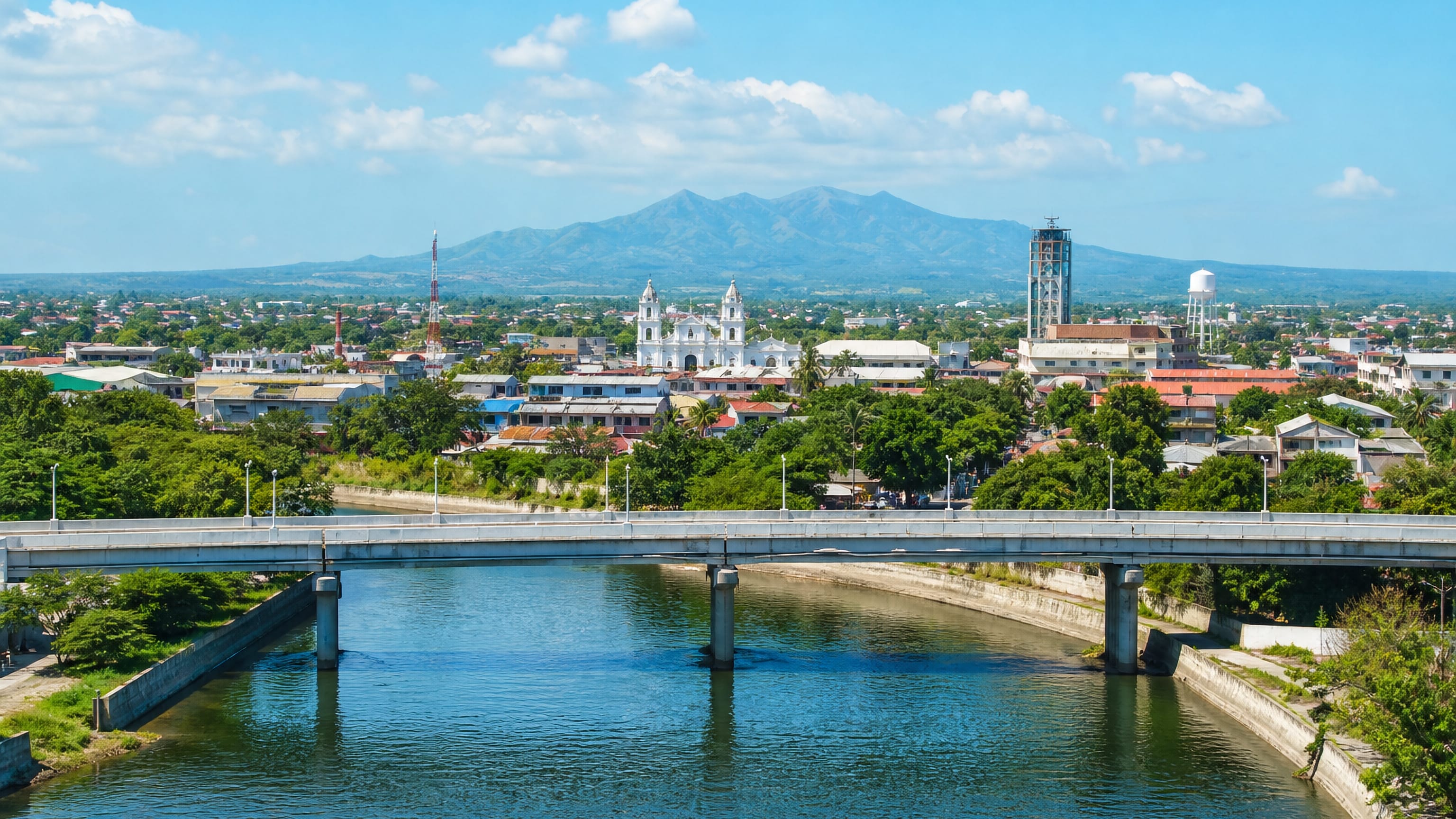 A river bridge leading into Mabalacat City, Pampanga, with a twin-towered church and mountains beyond.