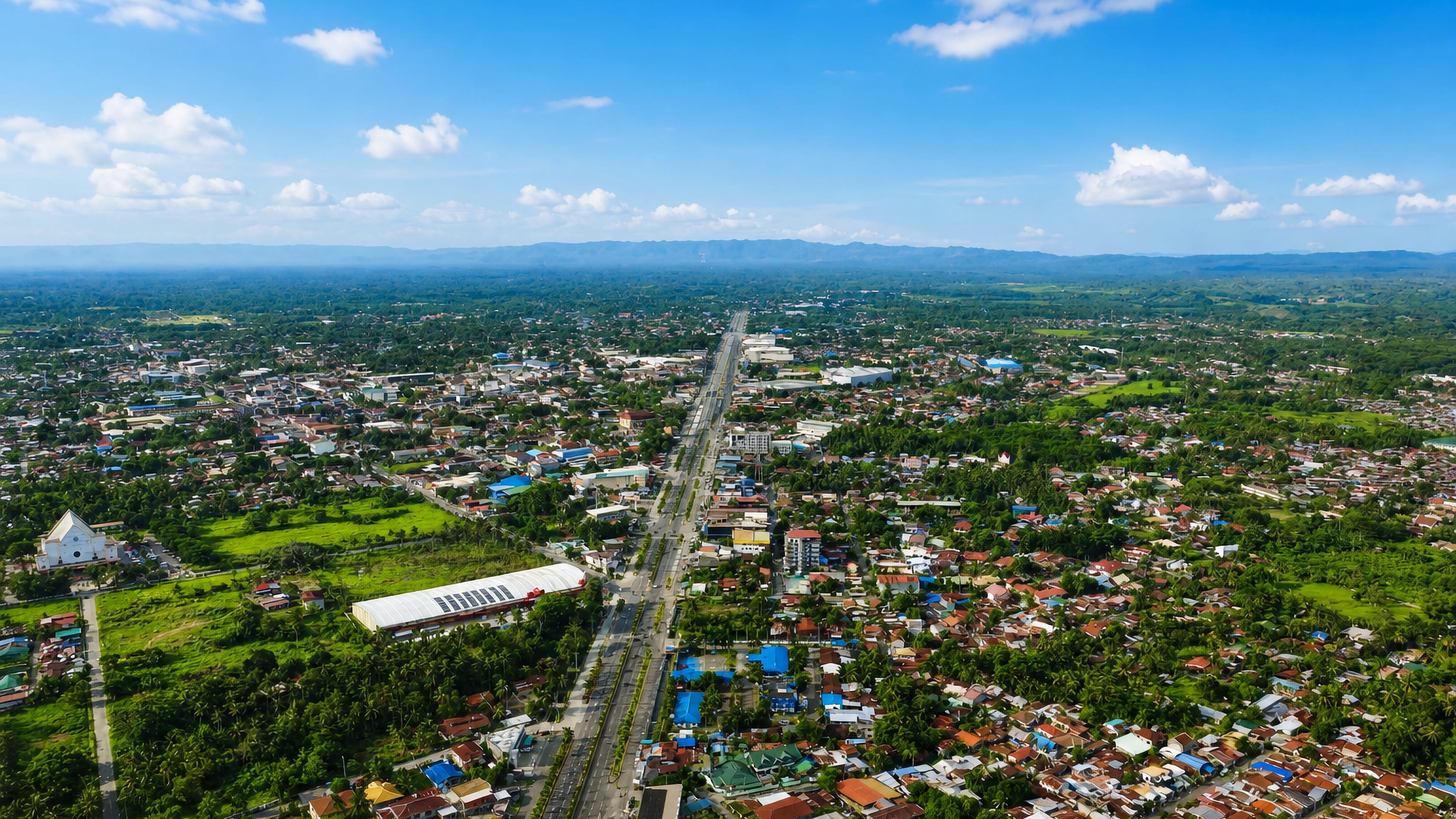 Aerial view of Magugpo Poblacion, Tagum, with its main highway, church and low rooftops below distant mountains.