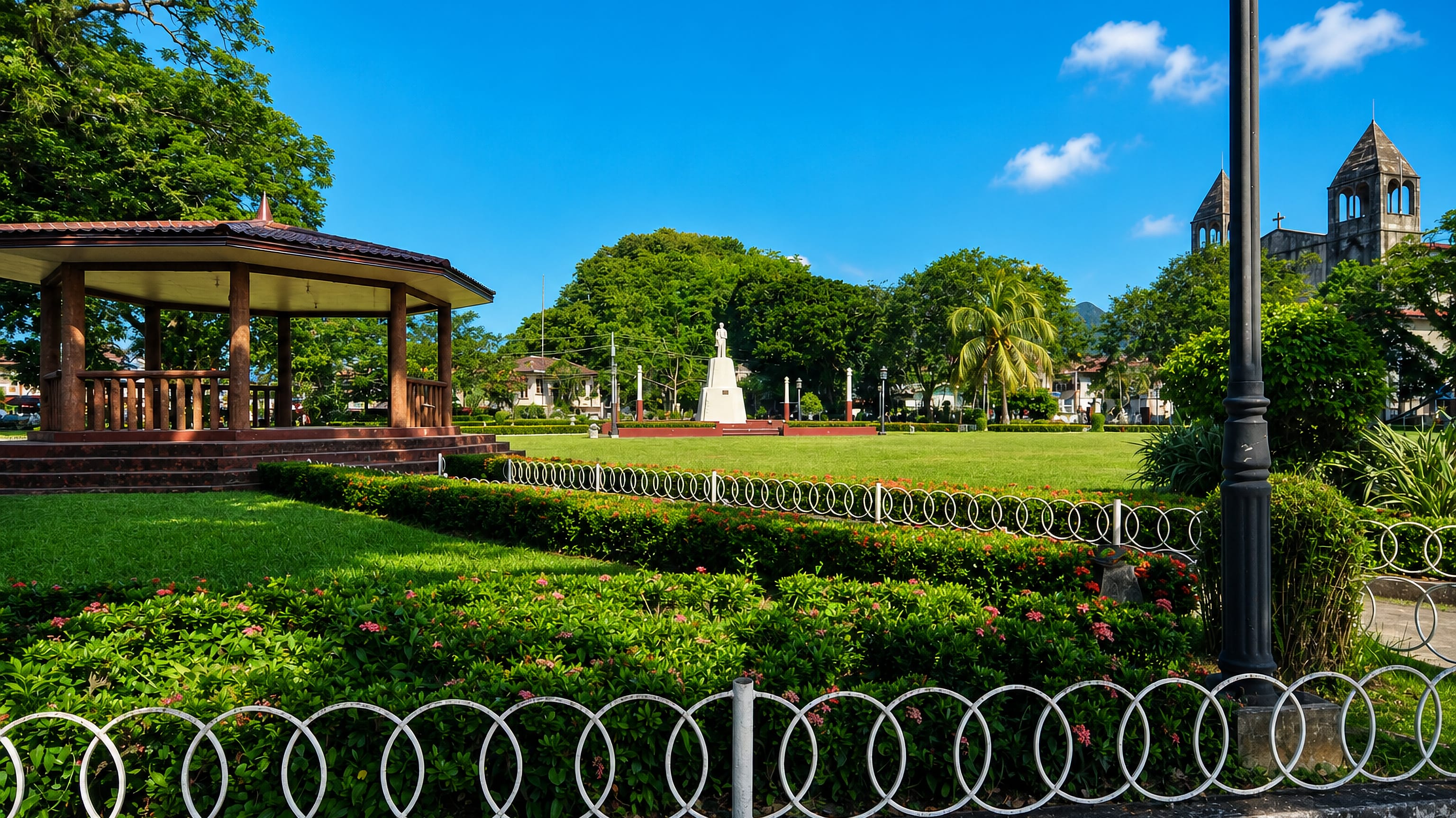 Dapitan's central plaza in the Philippines, with a wooden bandstand, a white monument and stone church towers.