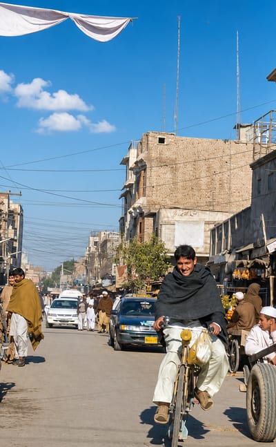 Busy daytime bazaar street in Bannu, Pakistan, with brick shopfronts, cyclists, market stalls and a donkey cart.