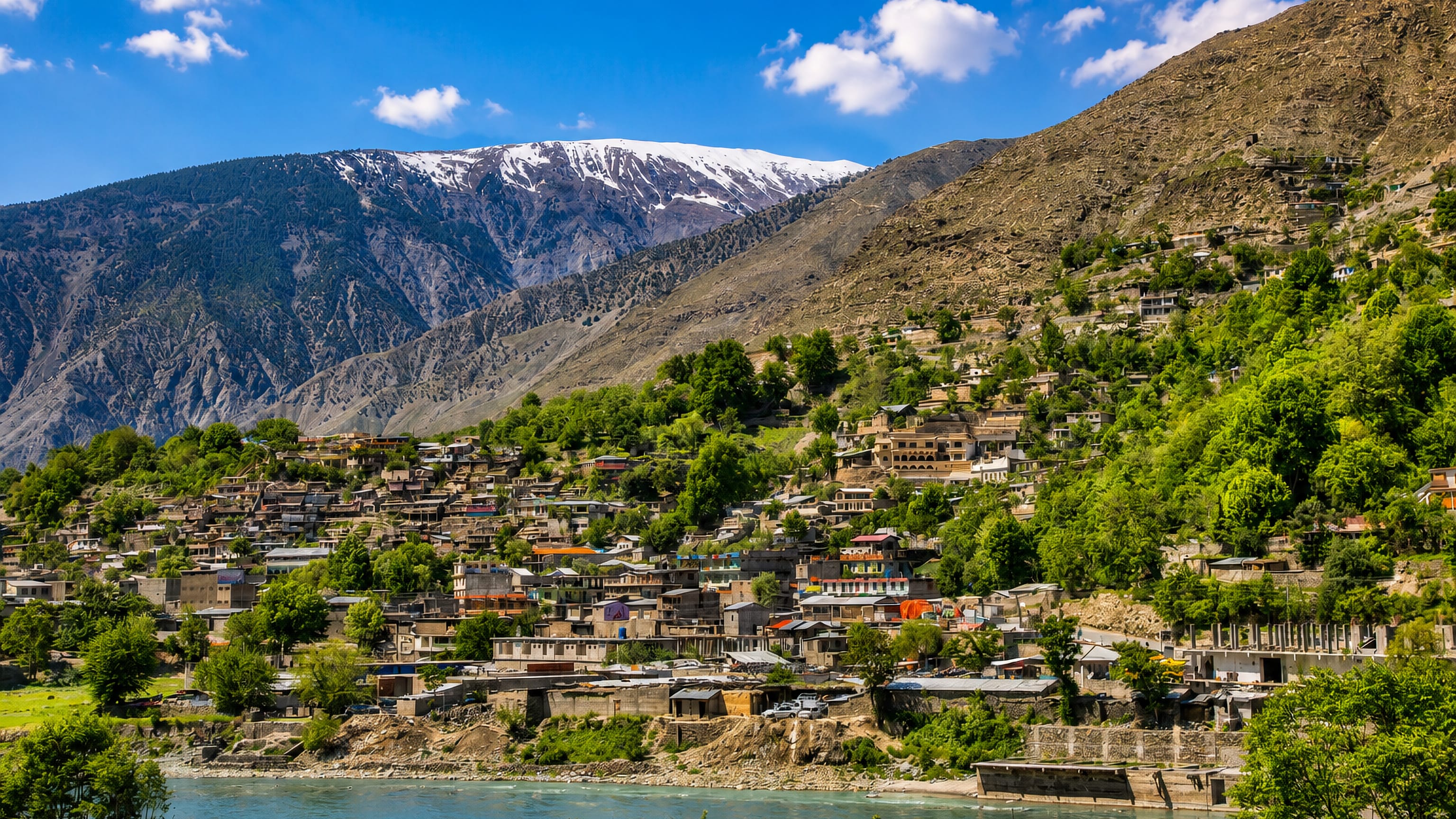 Hillside houses above the turquoise Chitral River in Chitral, Pakistan, below a snow-capped Hindu Kush ridge