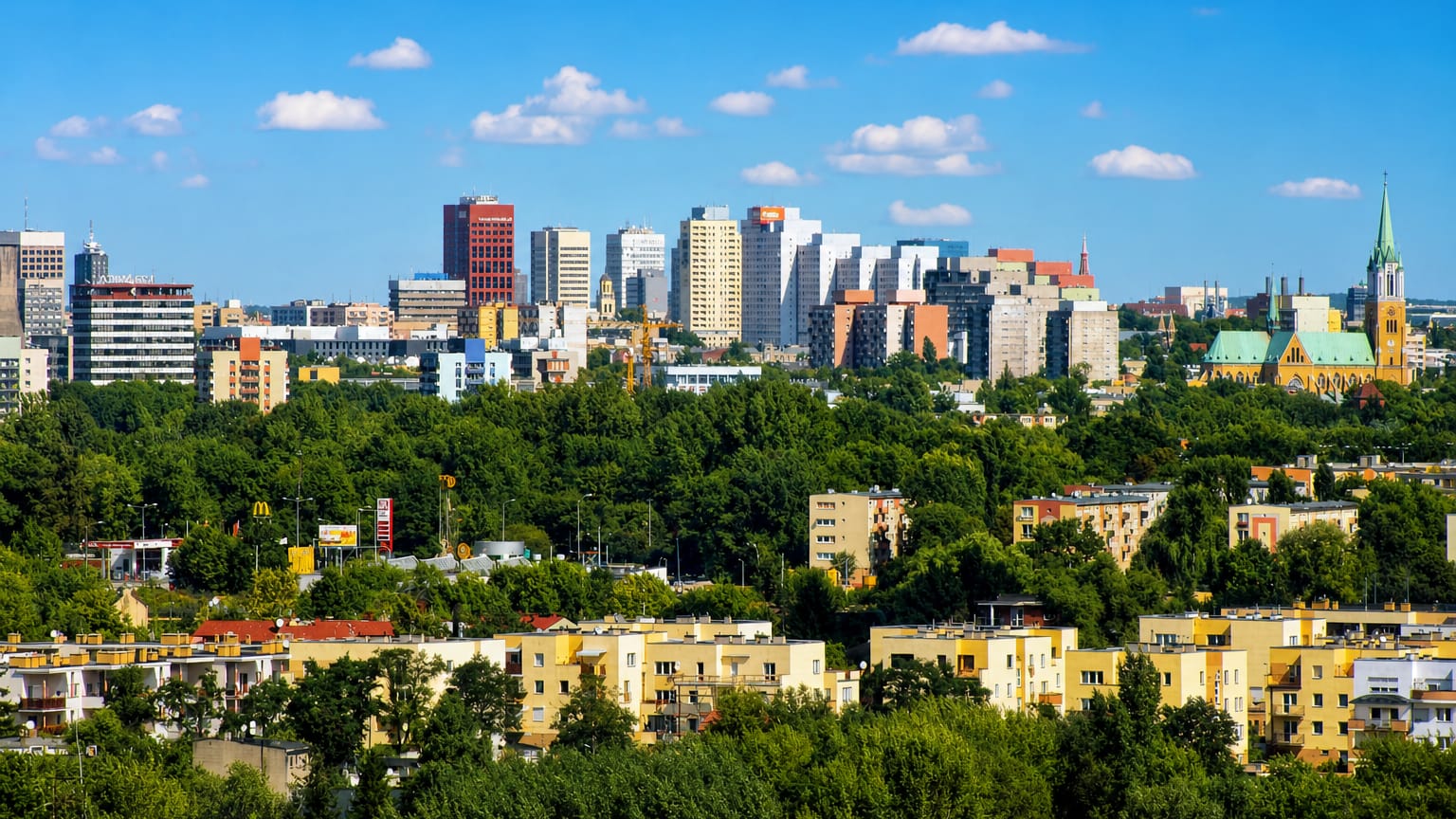 Łódź skyline with modern high-rises and a neo-Gothic cathedral spire rising above green trees and apartment blocks