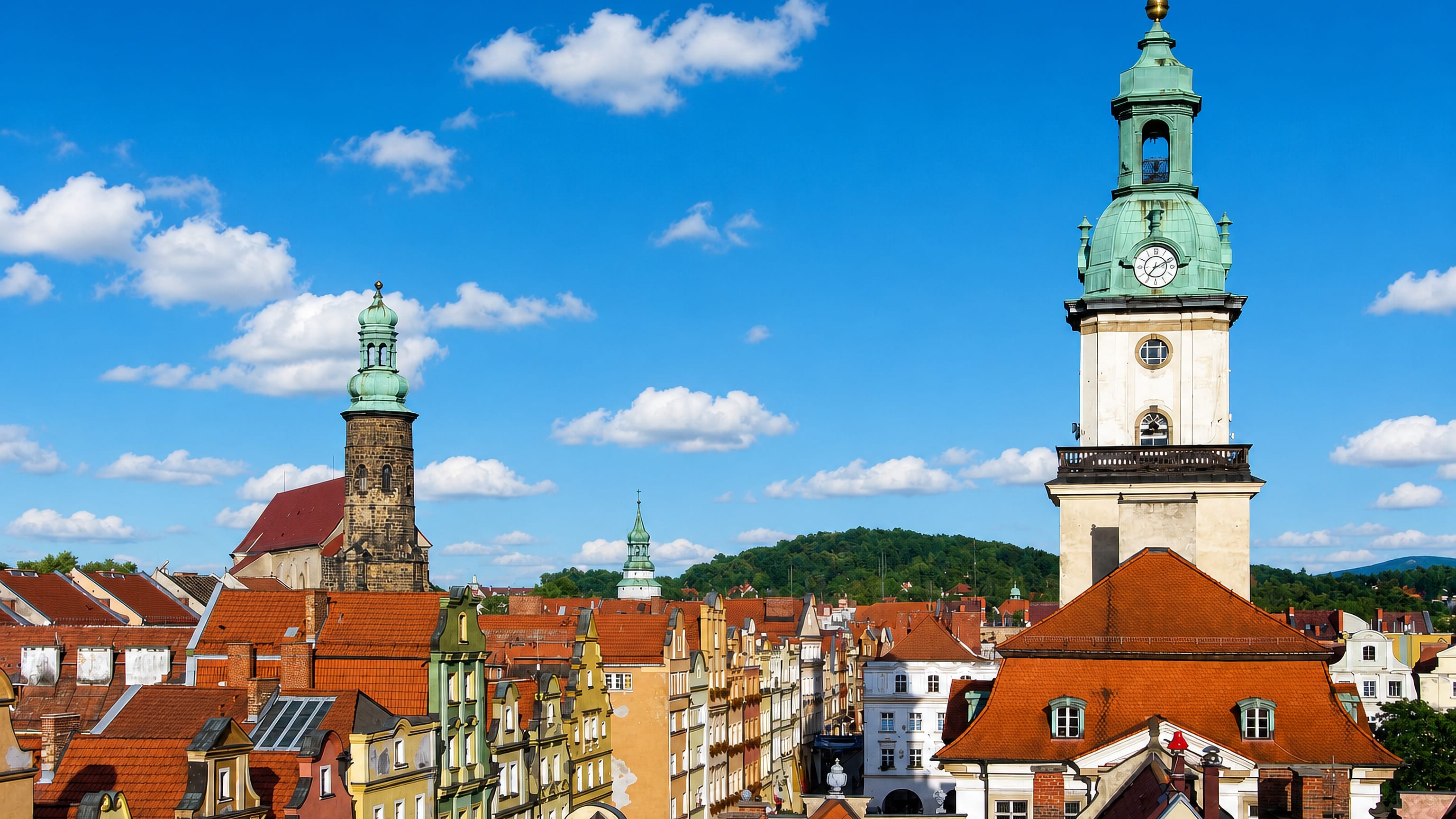 Red-tiled rooftops of Jelenia Gora's old town below the town hall clock tower and church spire, hills beyond.