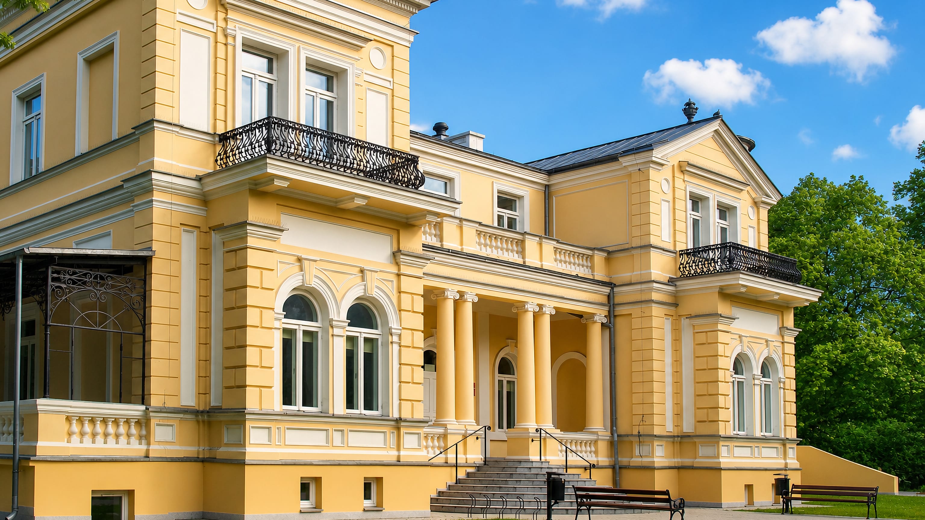 Restored yellow neoclassical palace with columned portico and wrought-iron balconies in Pruszków, Poland.