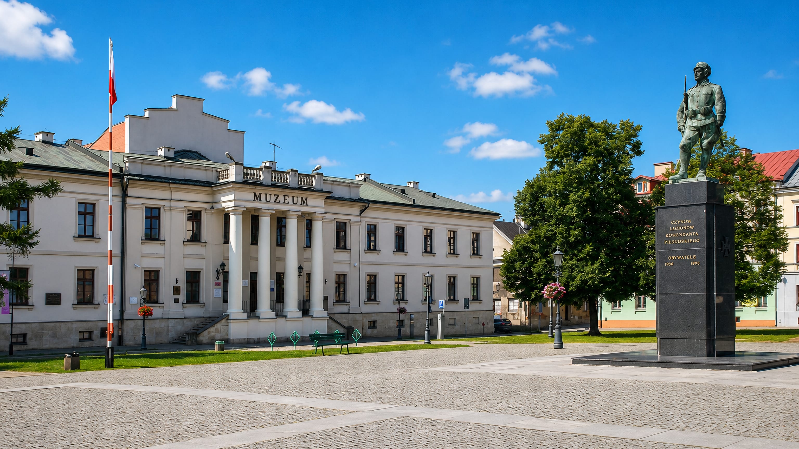 Radom's cobbled square with the Legions Monument and the neoclassical museum building under blue sky.