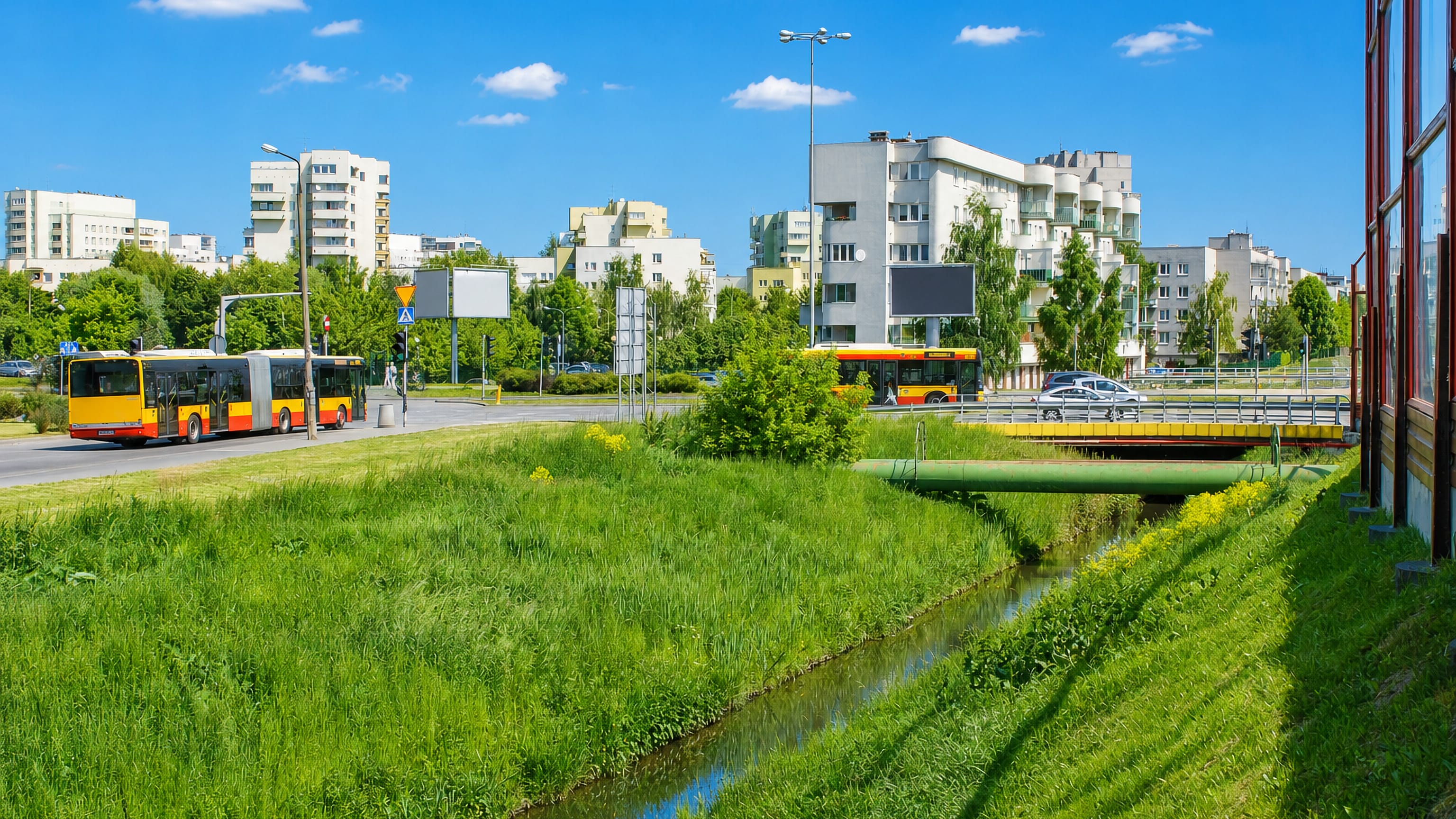 Articulated Warsaw buses passing pale apartment blocks beside a grassy canal in Goclaw, Warsaw.