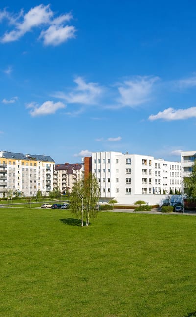Modern white and yellow apartment blocks behind a green park in Warsaw's Kabaty district under a blue sky.